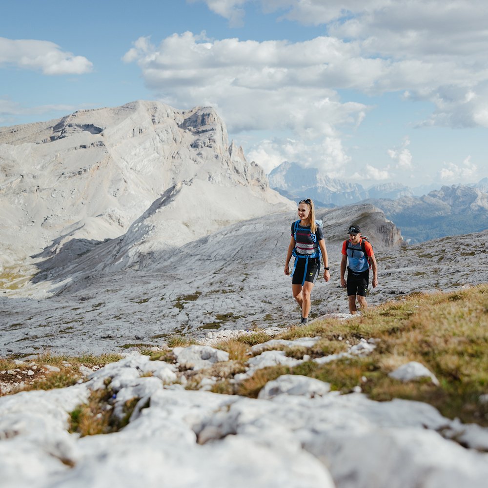 Two smiling hikers walk on a rocky path in Alta Badia, under a blue sky with clouds, with majestic Dolomites in the background. Ideal for outdoor activities.