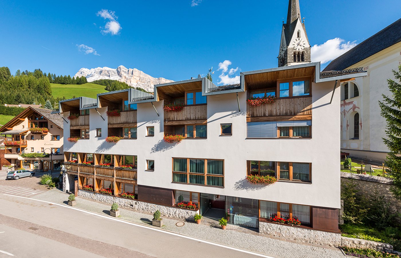 Facciata esterna dell'Hotel Cavallino a San Leonardo in Badia, con balconi in legno e vista sulle Dolomiti e sulla chiesa.
